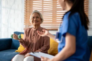 Smiling elderly woman lifting a small dumbbell at home while engaging in physical therapy with a caregiver.