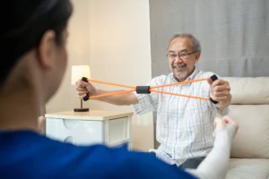 Elderly man doing stroke rehabilitation exercises with resistance band at home under caregiver guidance.